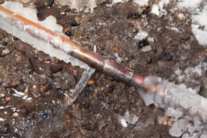 A 3/4" copper water main has frozen and cracked below ground outside a house. This is the line which runs from the street and services and individual house. In extreme cold this pipe can freeze and burst causing costly repairs. The background is excavated dirt.